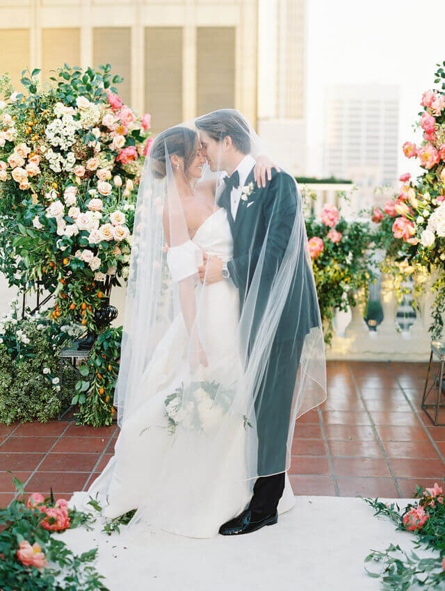Bride and groom beneath her veil beside a rose arch