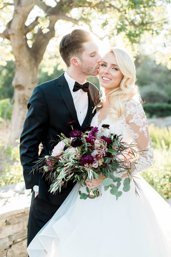 Groom kissing his bride at golden hour, burgundy bouquet in hand